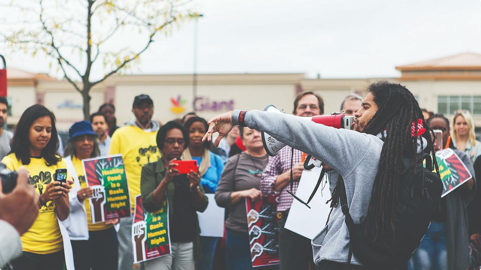 A public gathering outside a grocery store, where a speaker addresses community members with a megaphone while attendees hold signs advocating for healthy food